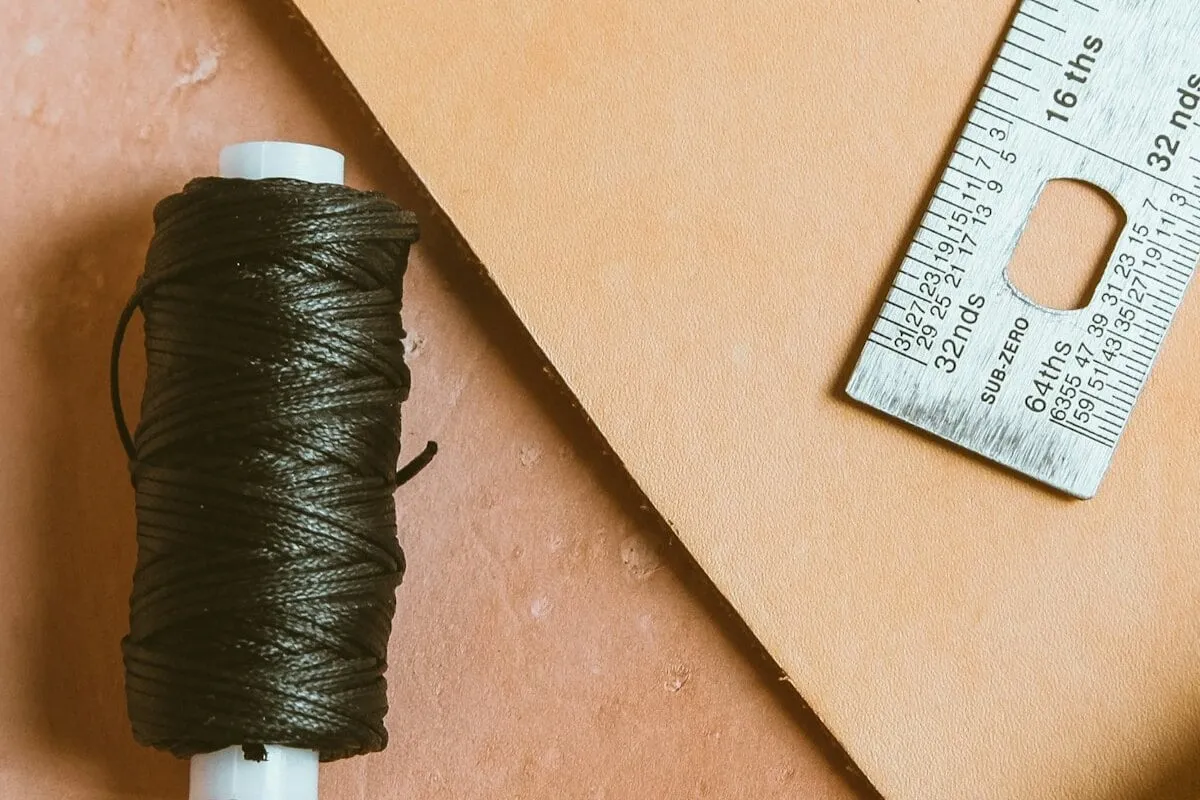 A spool of waxed linen thread beside a steel ruler on a sheet of leather.